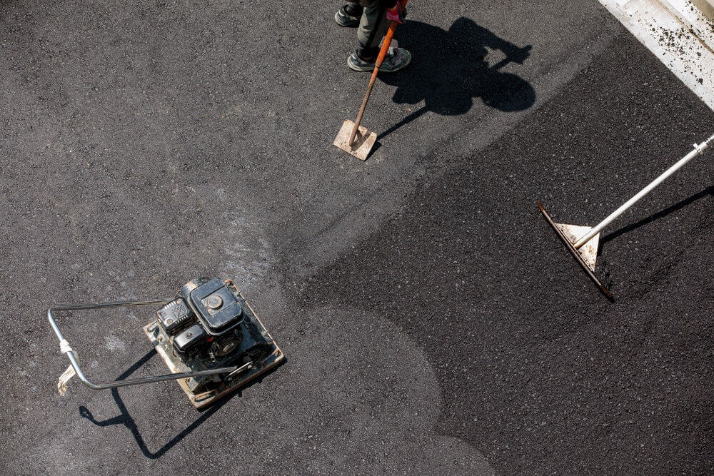 Worker compacting asphalt with vibrating plate and rake on freshly paved surface, showcasing precision akin to commercial roofing in Bethesda during exterior renovations.