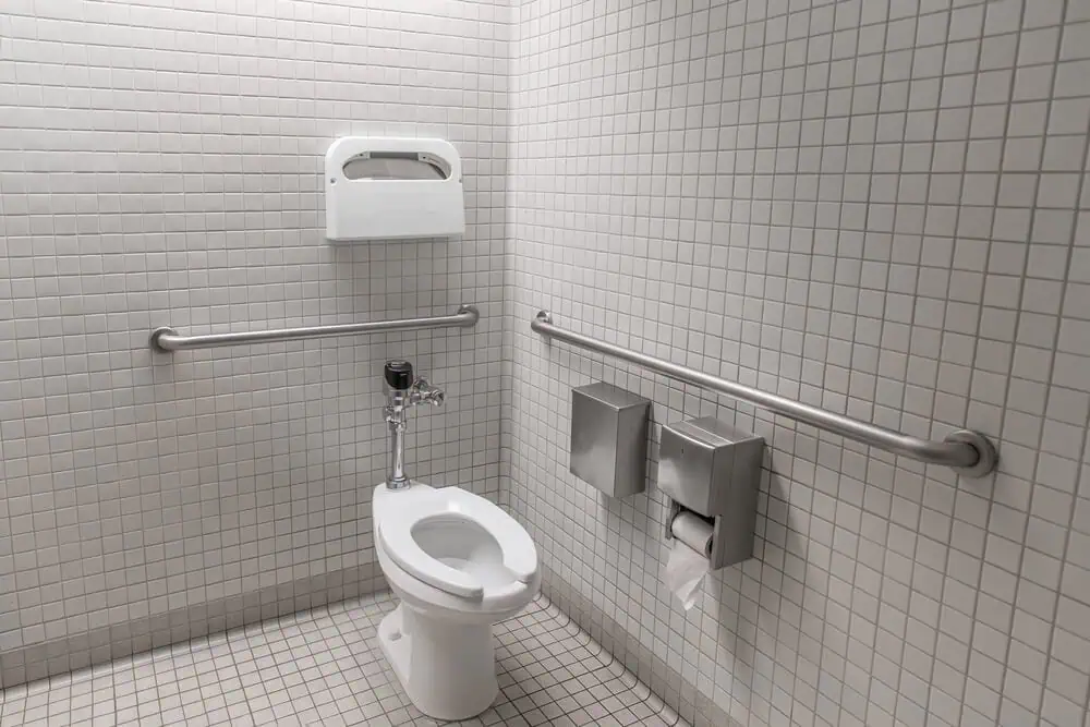 ADA-compliant commercial bathroom with accessible restroom, white tiled walls, handrails, toilet, and paper towel holder.