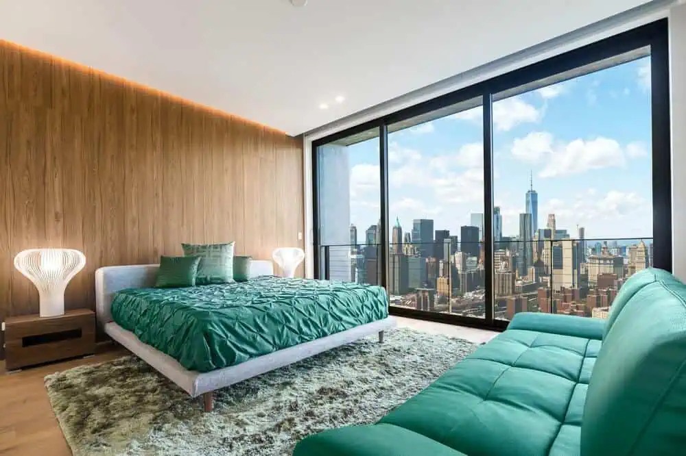 Modern bedroom with green bedding and sofa, floor-to-ceiling windows reveal city skyline; wood-paneled walls, plush rug add warmth.