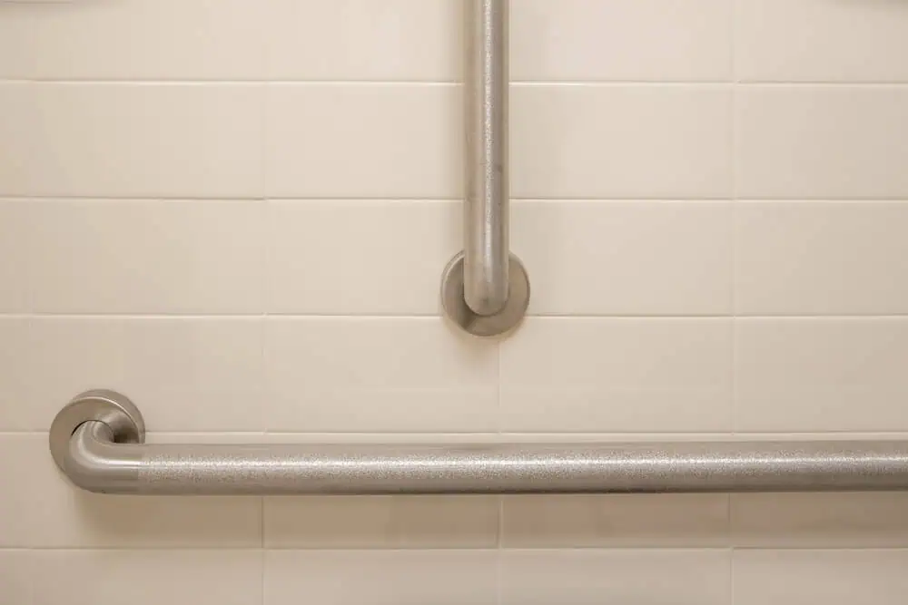 ADA-compliant metal grab bars on cream tiled bathroom wall, positioned horizontally and vertically for accessibility in commercial spaces.