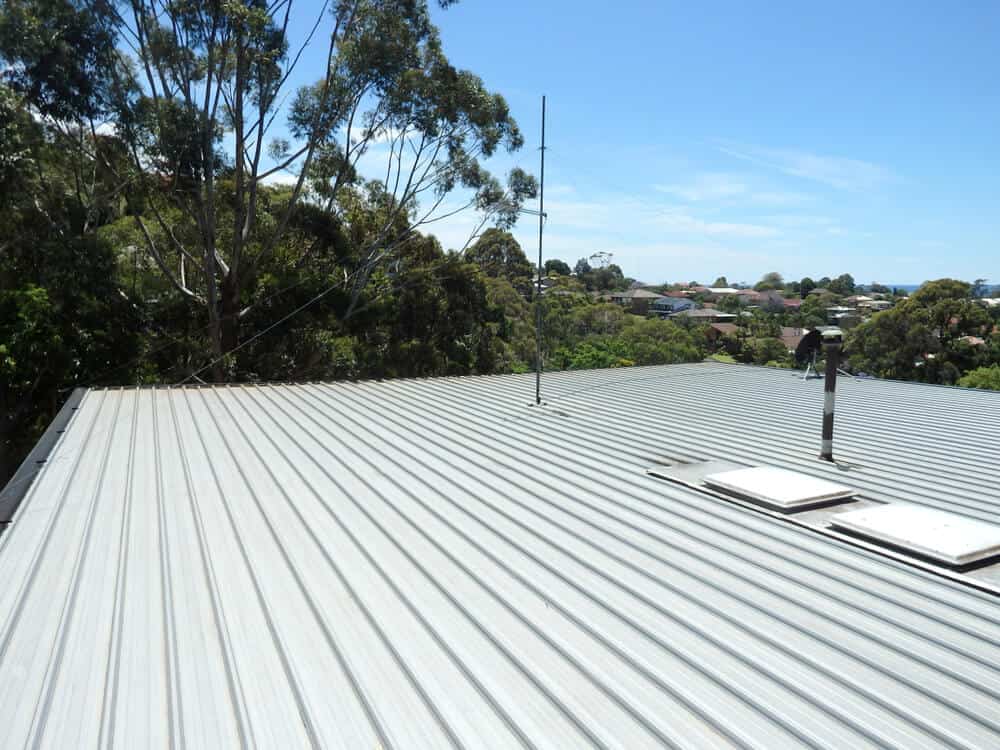 Commercial flat metal rooftop with skylights, vent, and antenna; surrounded by tall trees under a clear blue sky.