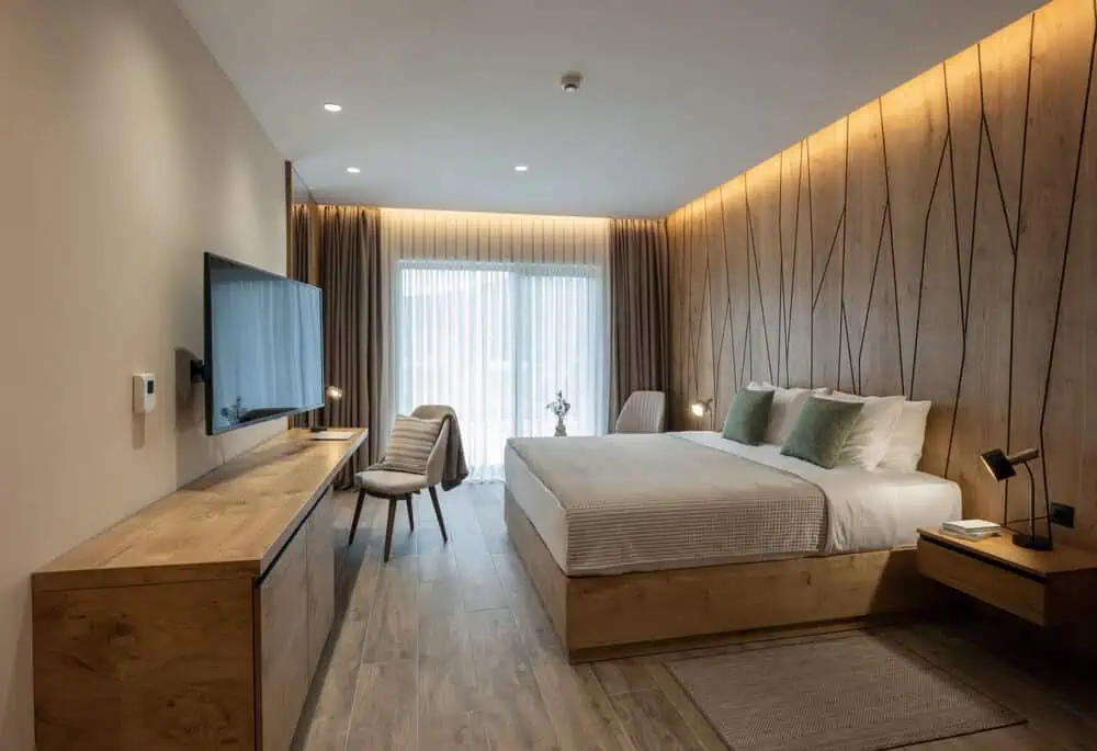 Modern hotel room interior with large bed, wooden accents, wall-mounted TV, desk and chair, window with curtains in a stylish renovation.