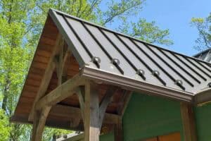 Green building with metal roof and wooden beams, featuring aligned snow guards; lush trees and clear blue sky in the background, perfect exterior renovation inspiration.