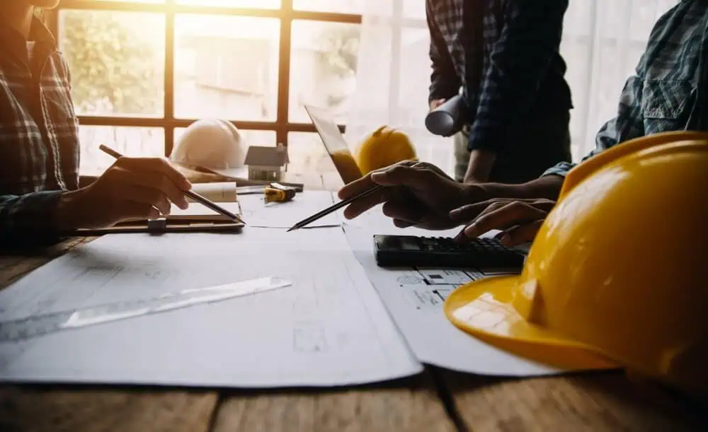 Engineers reviewing hotel renovation blueprints with pencils and calculator at table, yellow safety helmets symbolizing contractors, large window in background provides natural light.