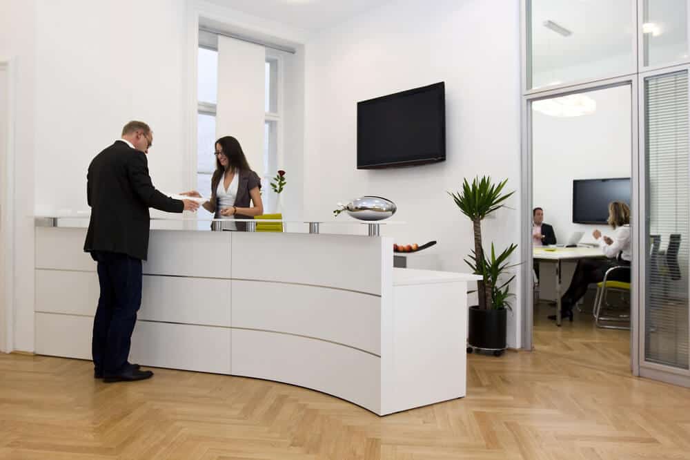 Man speaking with receptionist at modern white desk about commercial drywall repair in Columbia, MD office; meeting visible through glass door enhances busy environment.