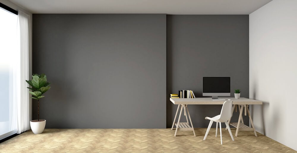 Minimalistic office design by commercial contractors in Bethesda, MD; features sleek desk, chair, computer, and books against gray wall. Potted plant near large window with white curtains. Interior renovation example.