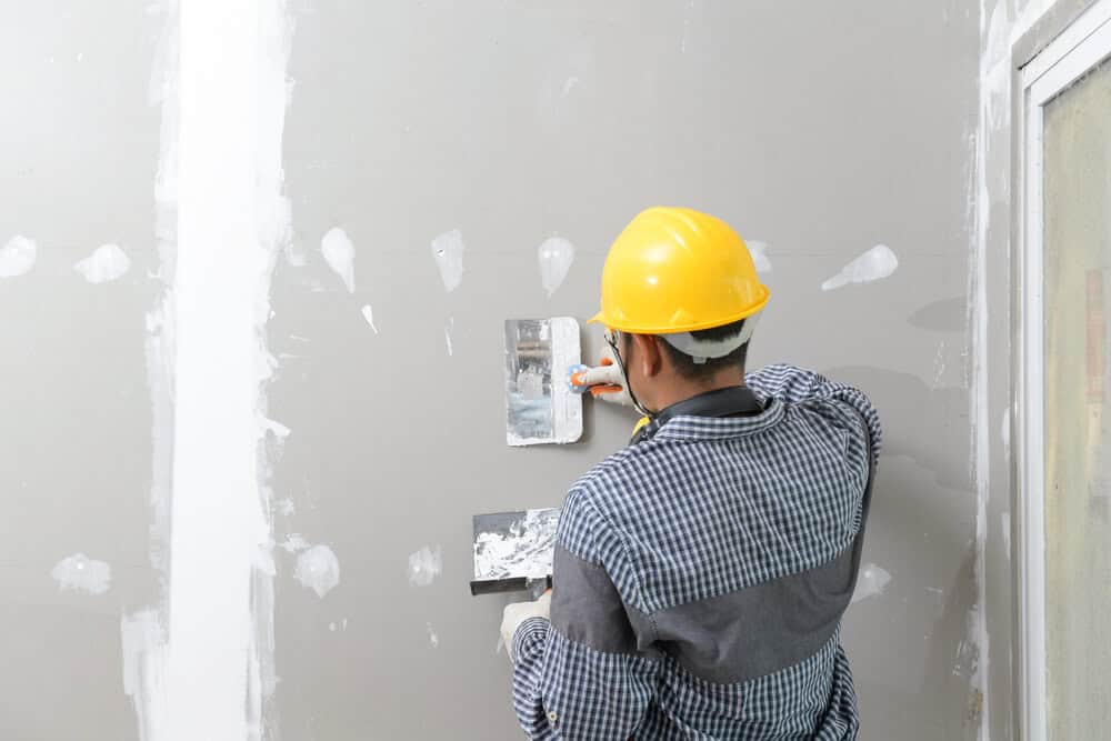 A worker in a checkered shirt and yellow hard hat skillfully applies plaster with a trowel, demonstrating expert commercial drywall repair & maintenance services in Columbia, MD.