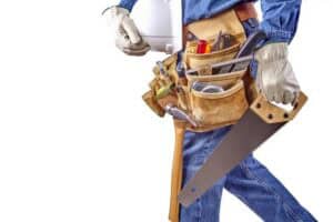Person in jeans and gloves holding toolbox and saw; tool belt organized with various renovation tools, perfect representation of professional exterior and interior construction services.