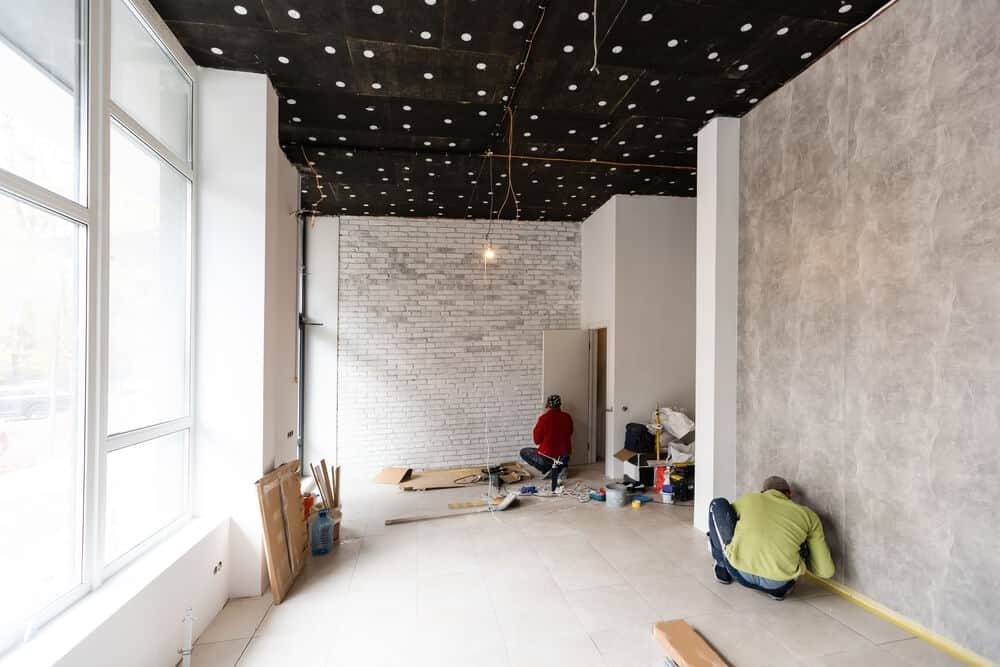 Two contractors working on interior renovations with tiled flooring, exposed brick walls, and black ceiling; tools and materials scattered for hotel remodeling project.