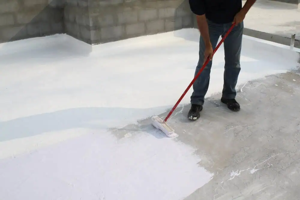 Worker applying white waterproof paint to commercial building floor with long-handled roller for even coat and enhanced durability in renovation project.