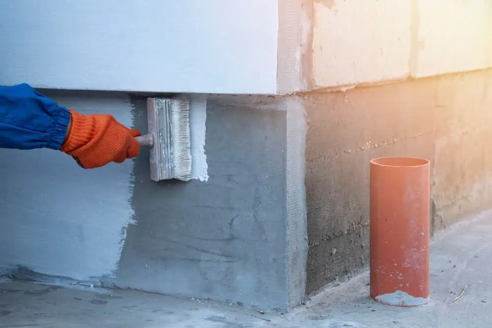 A worker in orange gloves painting a concrete wall corner white, emphasizing commercial building waterproofing; nearby is an orange pipe.