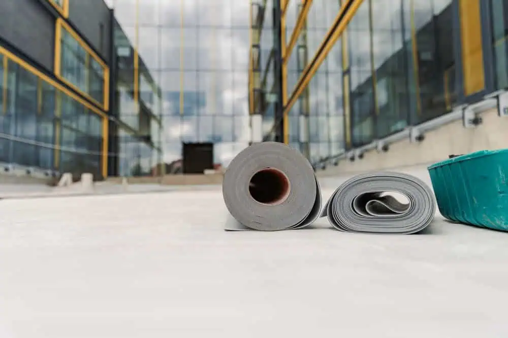 Two rolls of roofing material on concrete near a modern building with glass windows, highlighting commercial roof replacement in urban construction.