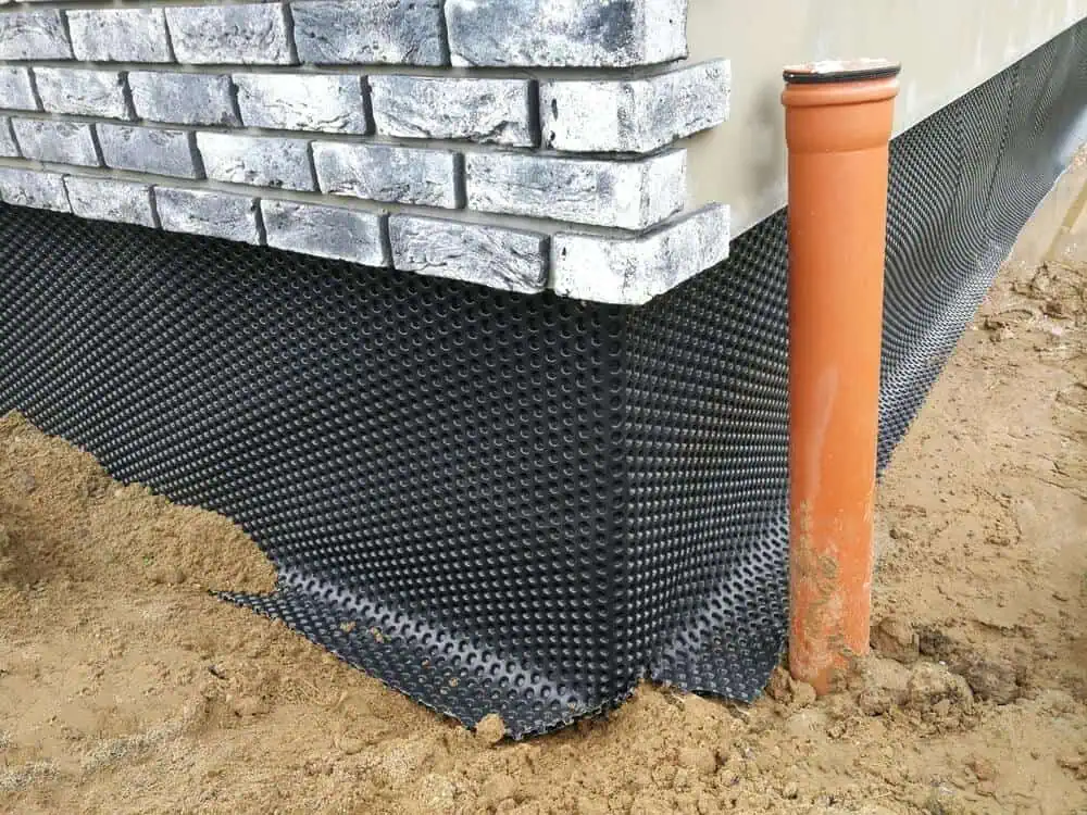 Close-up view of a building foundation under construction, showcasing commercial waterproofing methods with a dimpled membrane and orange drainage pipe against brick wall.