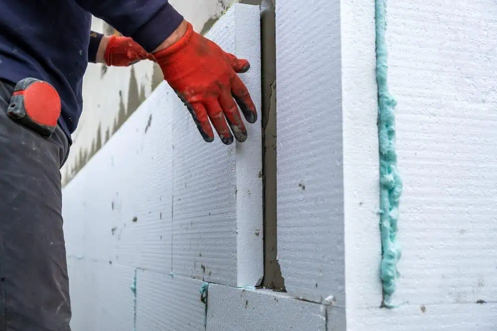 A worker in red gloves installs white foam insulation panels with visible green sealant for commercial facade renovation.