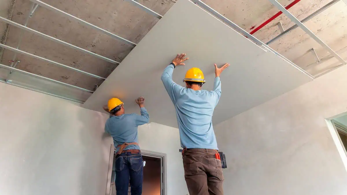 Two drywall installers work together to hang a drywall ceiling panel