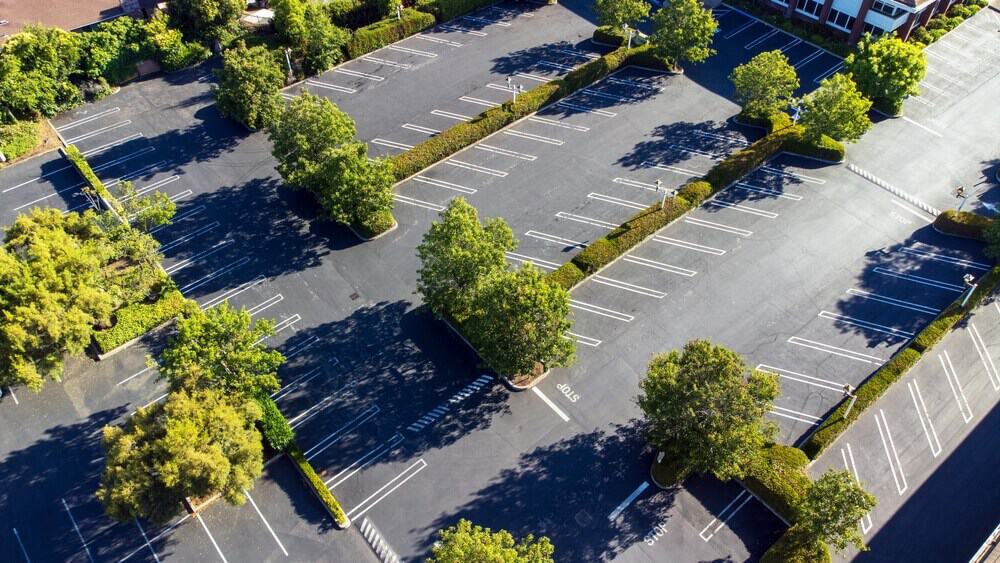 Aerial view of a spacious empty parking lot with multiple rows of parking spaces, encircled by lush trees and greenery, showcasing potential for construction and renovation projects.