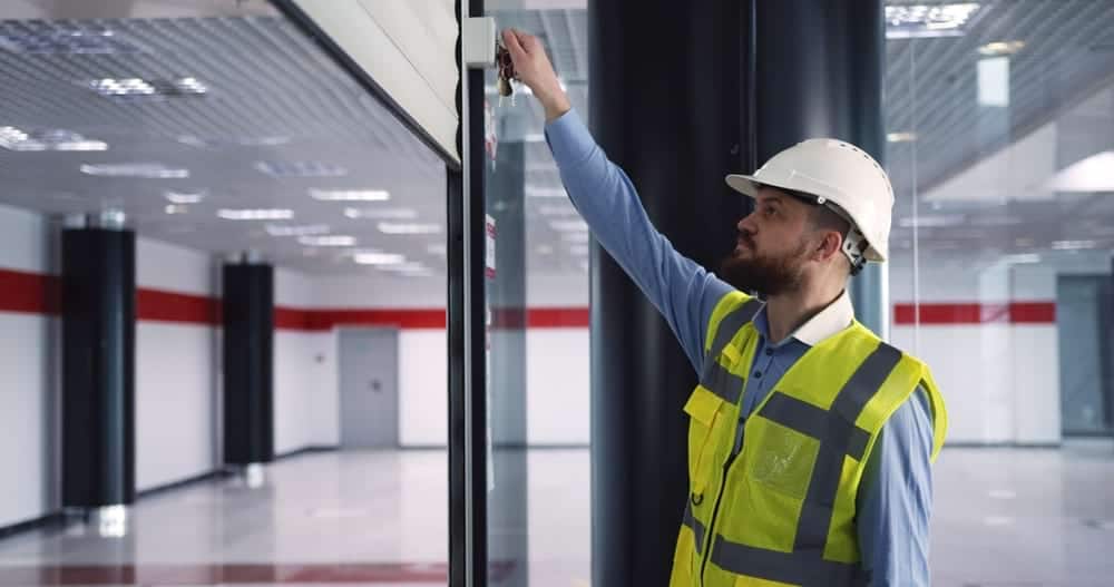 Construction worker in safety vest and hard hat adjusting device on glass wall during commercial door repair in modern building interior.