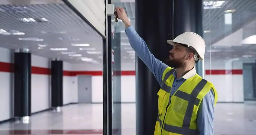 Construction worker in safety vest and hard hat adjusting device on glass wall during commercial door repair in modern building interior.