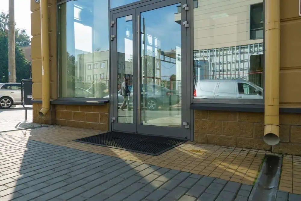 Modern glass entrance door with black floor mat, yellow walls, visible drainage pipes; commercial building exterior ideal for professional door repair and renovation services.