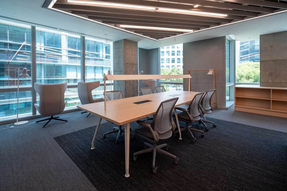 Modern office renovation featuring large wooden table, stylish chairs, lounge seating by floor-to-ceiling windows with city views; perfect for commercial interiors and professional door repair services.