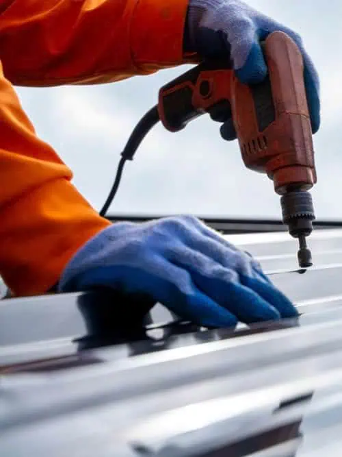 Worker in blue gloves and orange jacket using power drill to fasten screw into metal sheet, demonstrating professional commercial metal roofing installation.